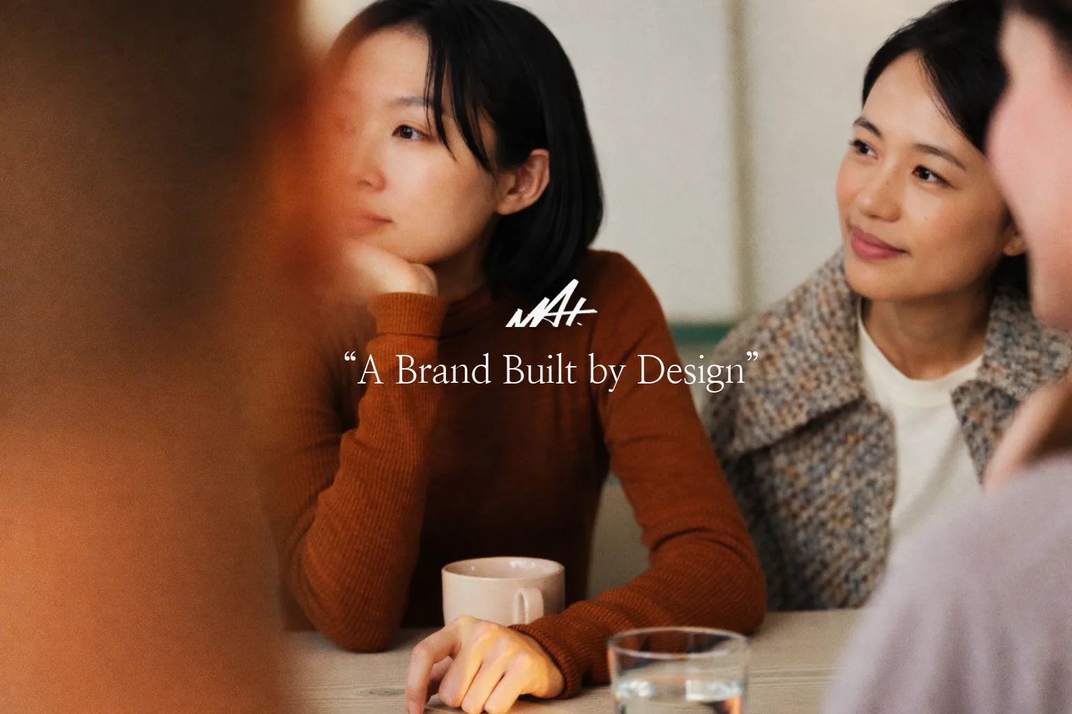 테이블에 앉은 두 인물 위에 MAT 로고와 ‘A Brand Built by Design’ 문구를 배치한 이미지